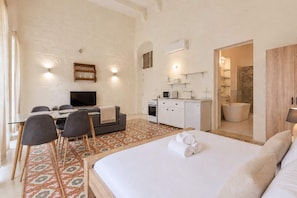 A hotel room with a bed, a TV, a dining table with chairs, a kitchenette, and a bathroom. - Historic Flat for 5 near St. John's Cathedral (Valletta)