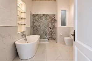 A bathroom with a freestanding bathtub, a toilet, and a shower area with patterned tiles. - Historic Flat for 5 near St. John's Cathedral (Valletta)