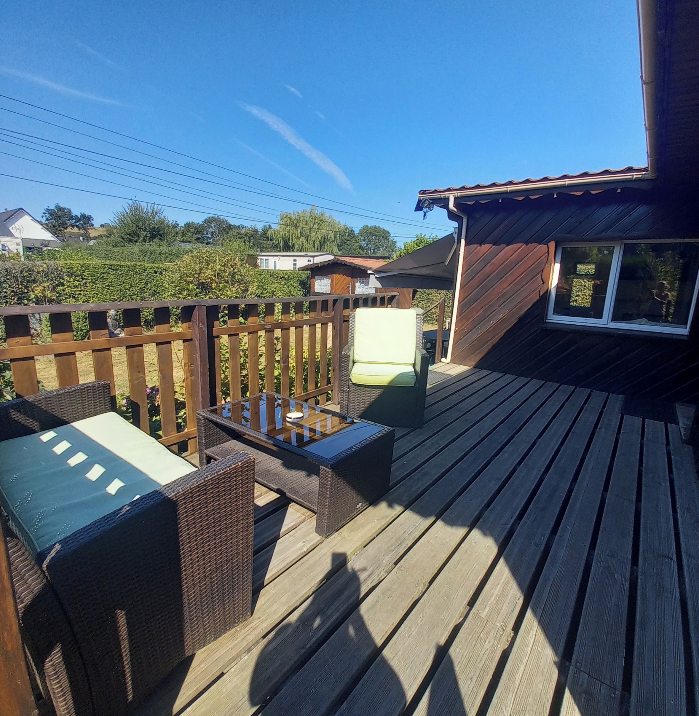 Een houten terras met buitenmeubilair, een klein gebouw met een raam en een haag op de achtergrond.