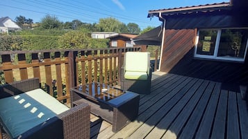 Een houten terras met buitenmeubilair, een klein gebouw met een raam en een haag op de achtergrond.