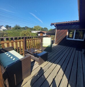 A wooden deck with outdoor furniture, a small building with a window, and a hedge in the background.