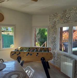 A room with a foosball table, two windows, and patterned wallpaper.