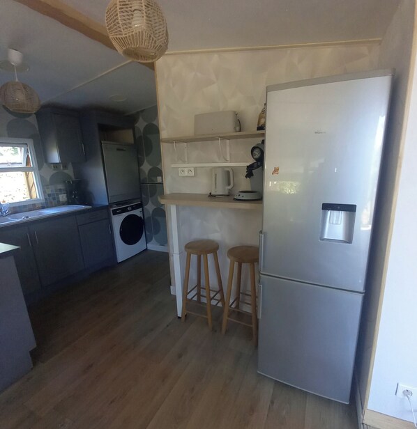 A compact kitchen with a built-in washing machine, a refrigerator, wooden stools, and a hanging light fixture.