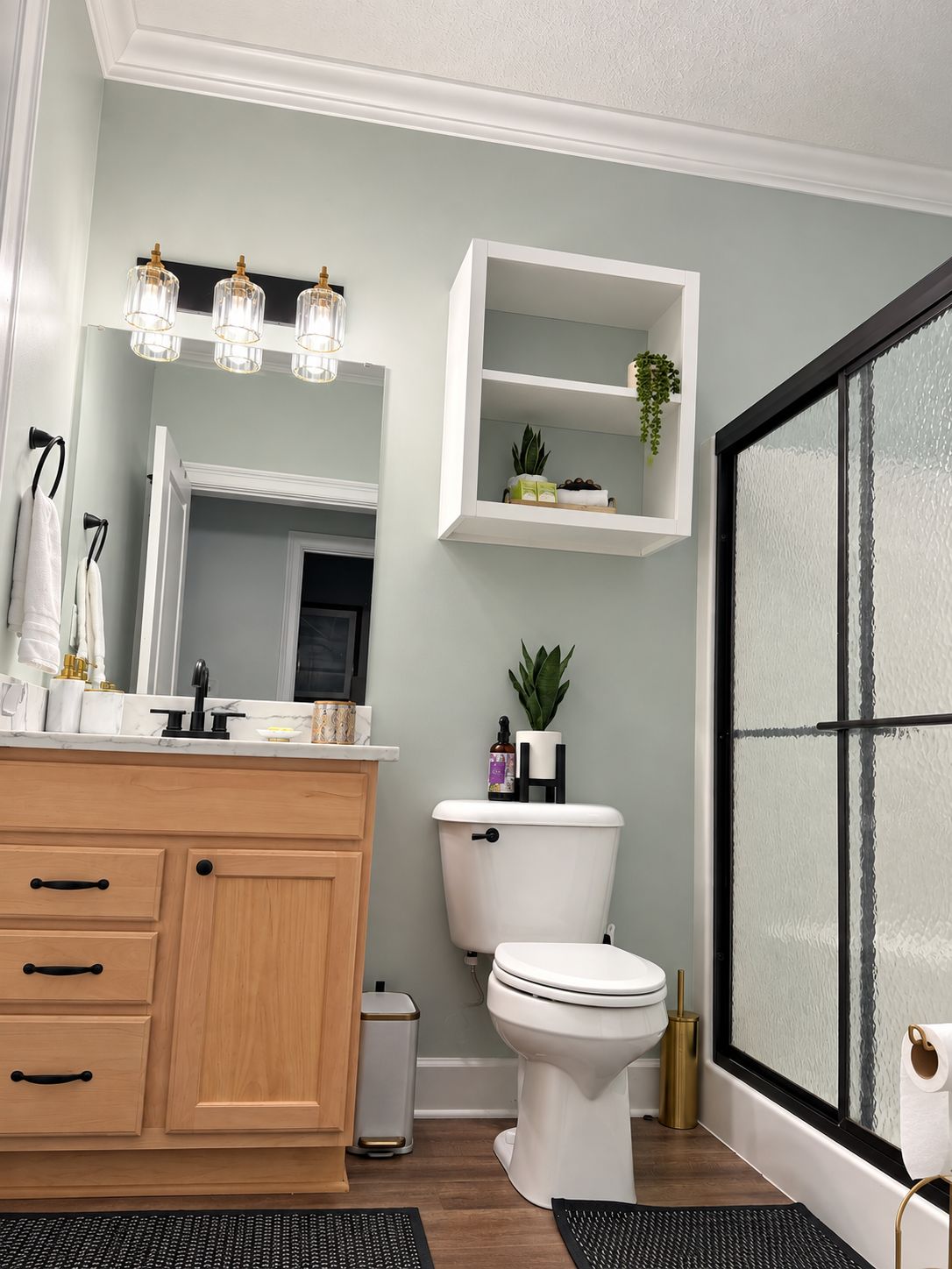 A modern bathroom with a wooden vanity, a toilet, a shelf with plants, and a frosted glass shower door.