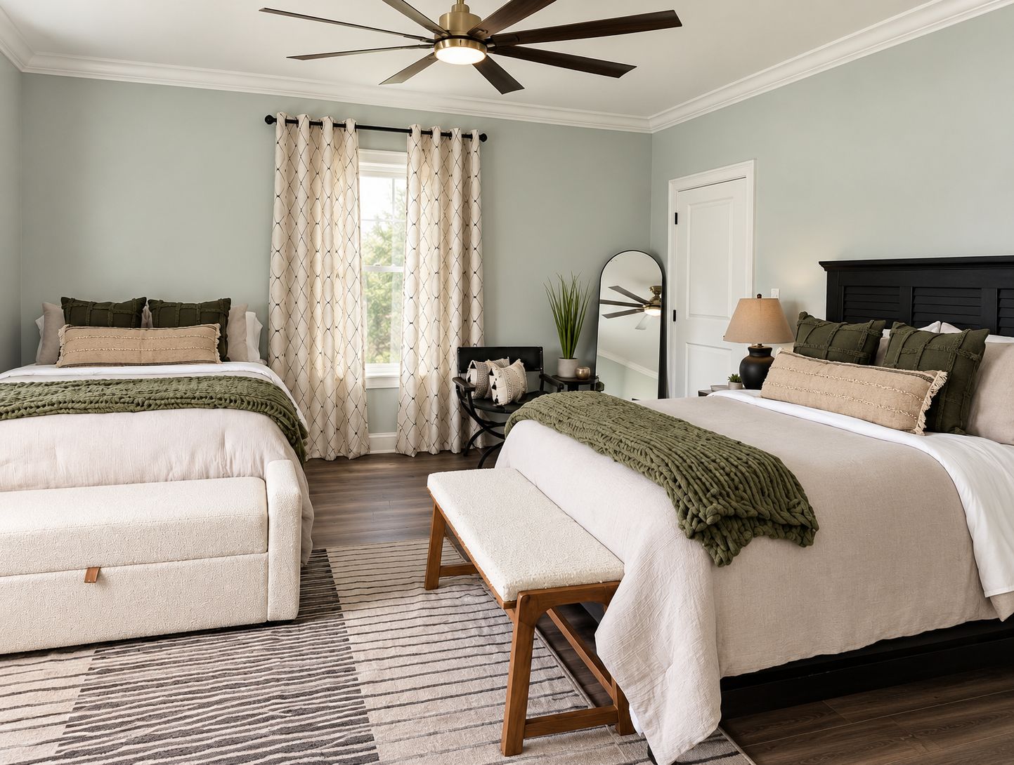 A bedroom with two beds, a ceiling fan, a window with curtains, a bedside table with a lamp, and a mirror.