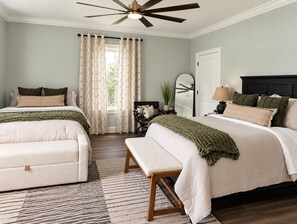 A bedroom with two beds, a ceiling fan, a window with curtains, a bedside table with a lamp, and a mirror.