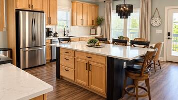 A modern kitchen with wooden cabinets, a stainless steel refrigerator, a large island with bar stools, and a pendant light.