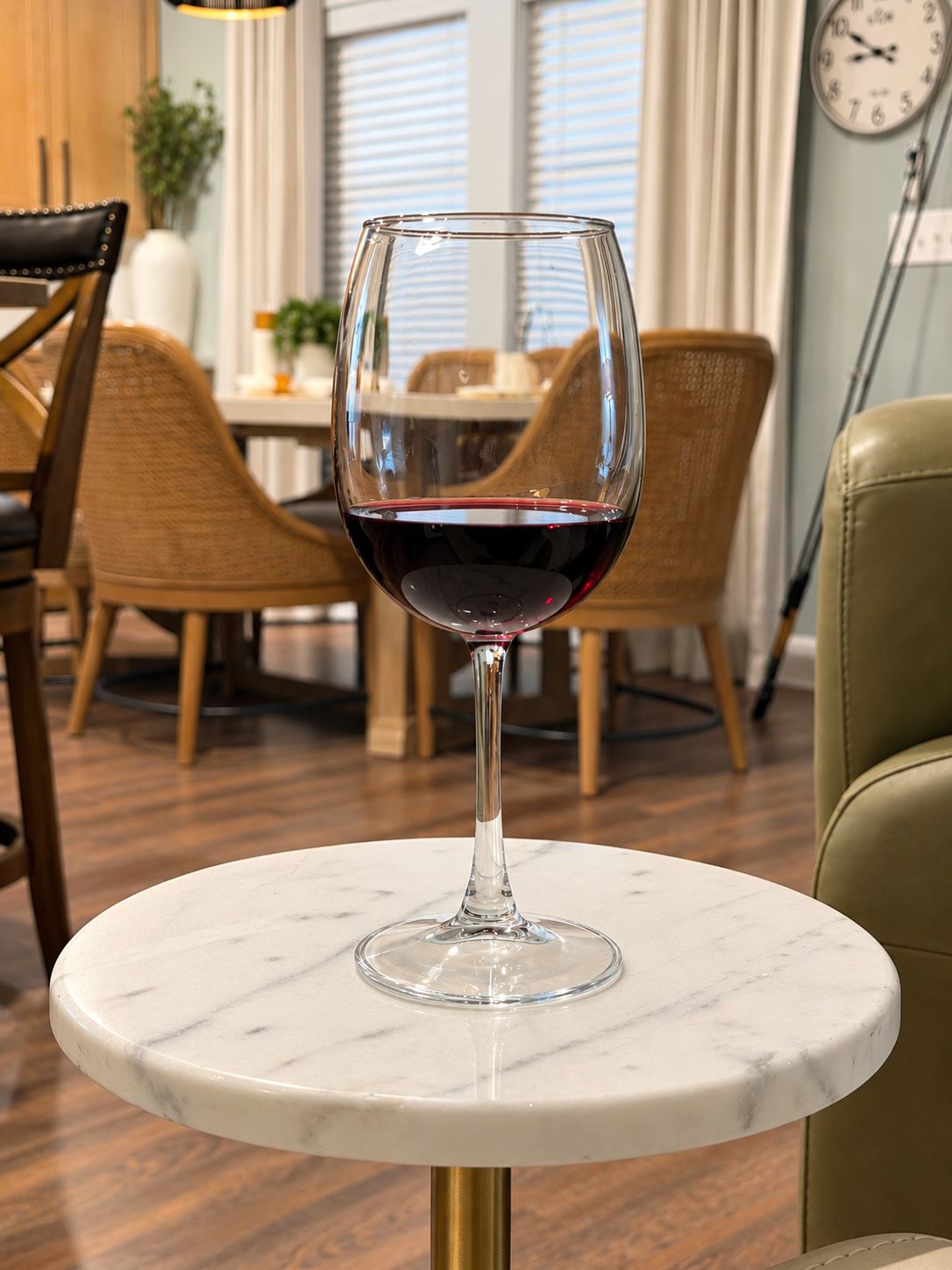 A wine glass on a marble table in a dining area with wicker chairs and a clock on the wall.