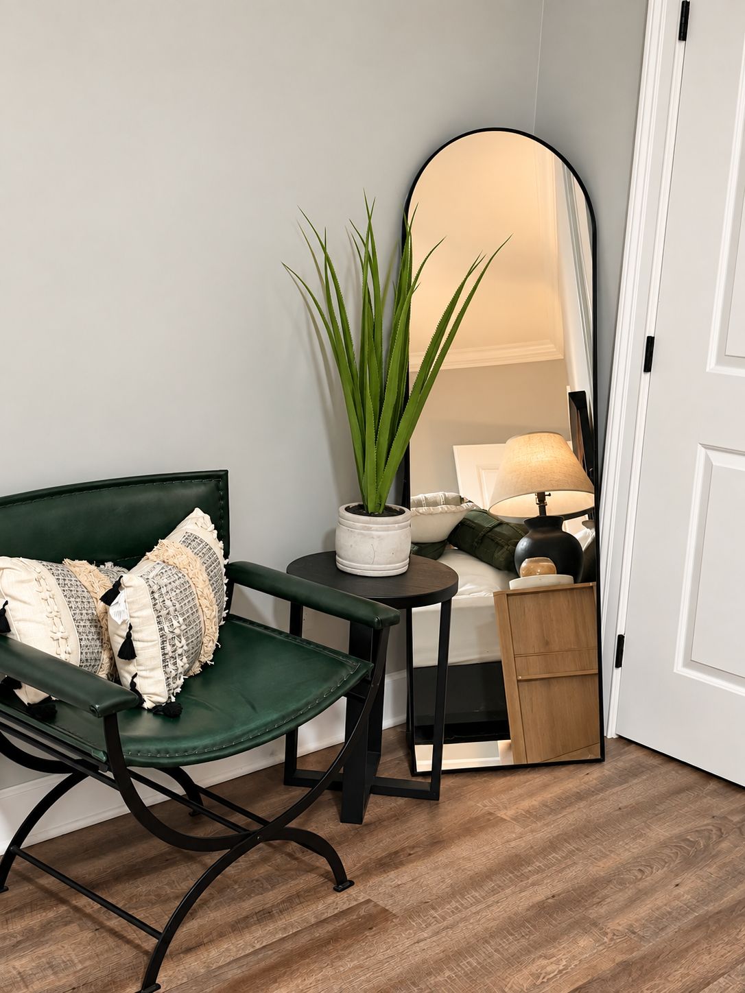 A green cushioned chair with patterned pillows, a small round table with a potted plant, a mirror reflecting a bed and lamp, and a wooden cabinet.