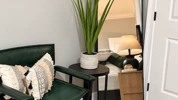A green cushioned chair with patterned pillows, a small round table with a potted plant, a mirror reflecting a bed and lamp, and a wooden cabinet.