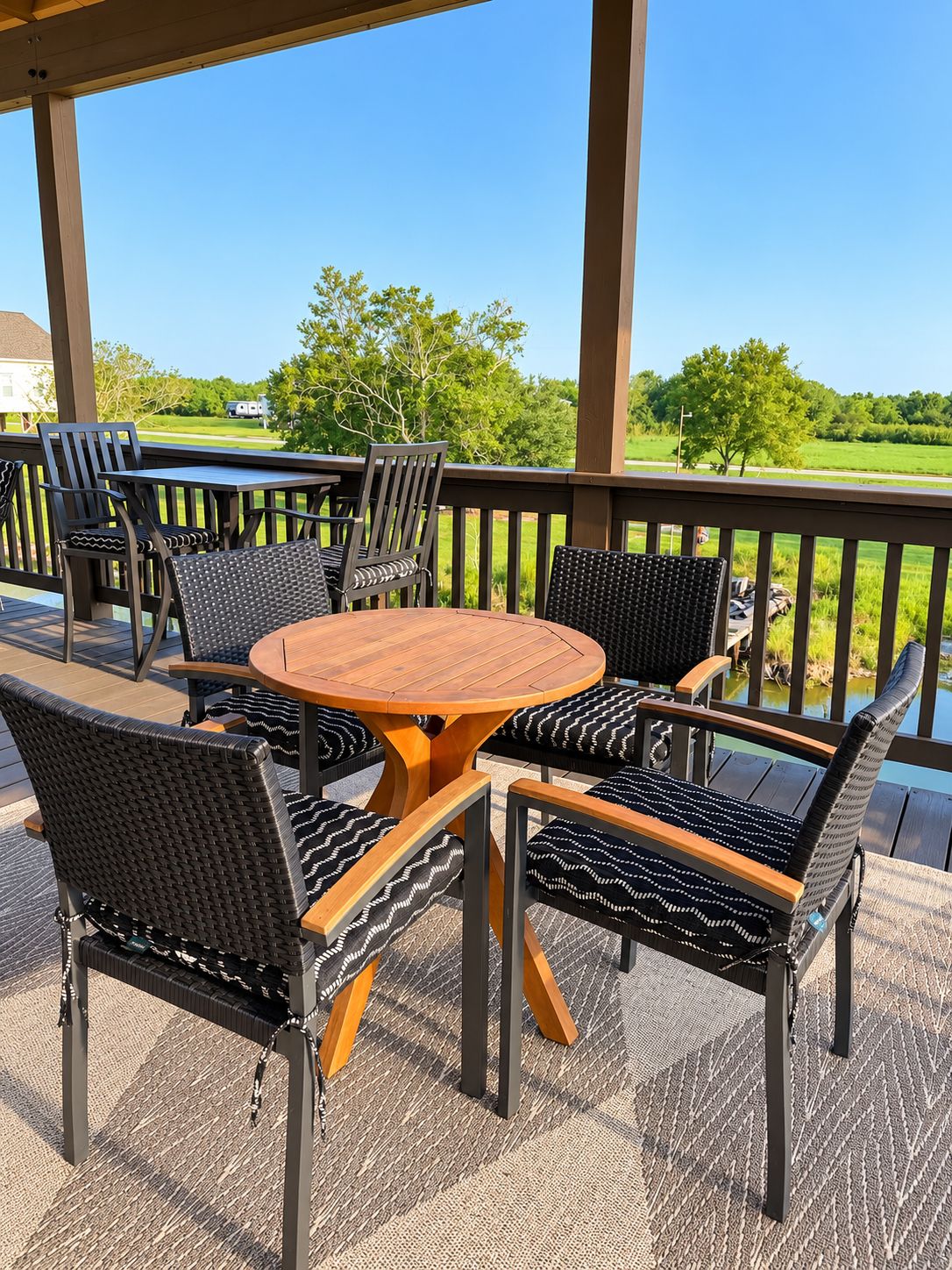 A covered outdoor seating area with a round table and chairs, a view of a green landscape, and a wooden railing.