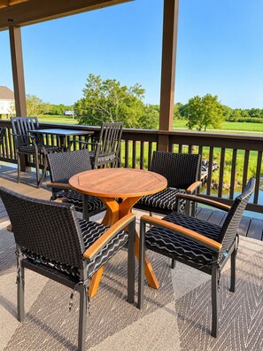 A covered outdoor seating area with a round table and chairs, a view of a green landscape, and a wooden railing.