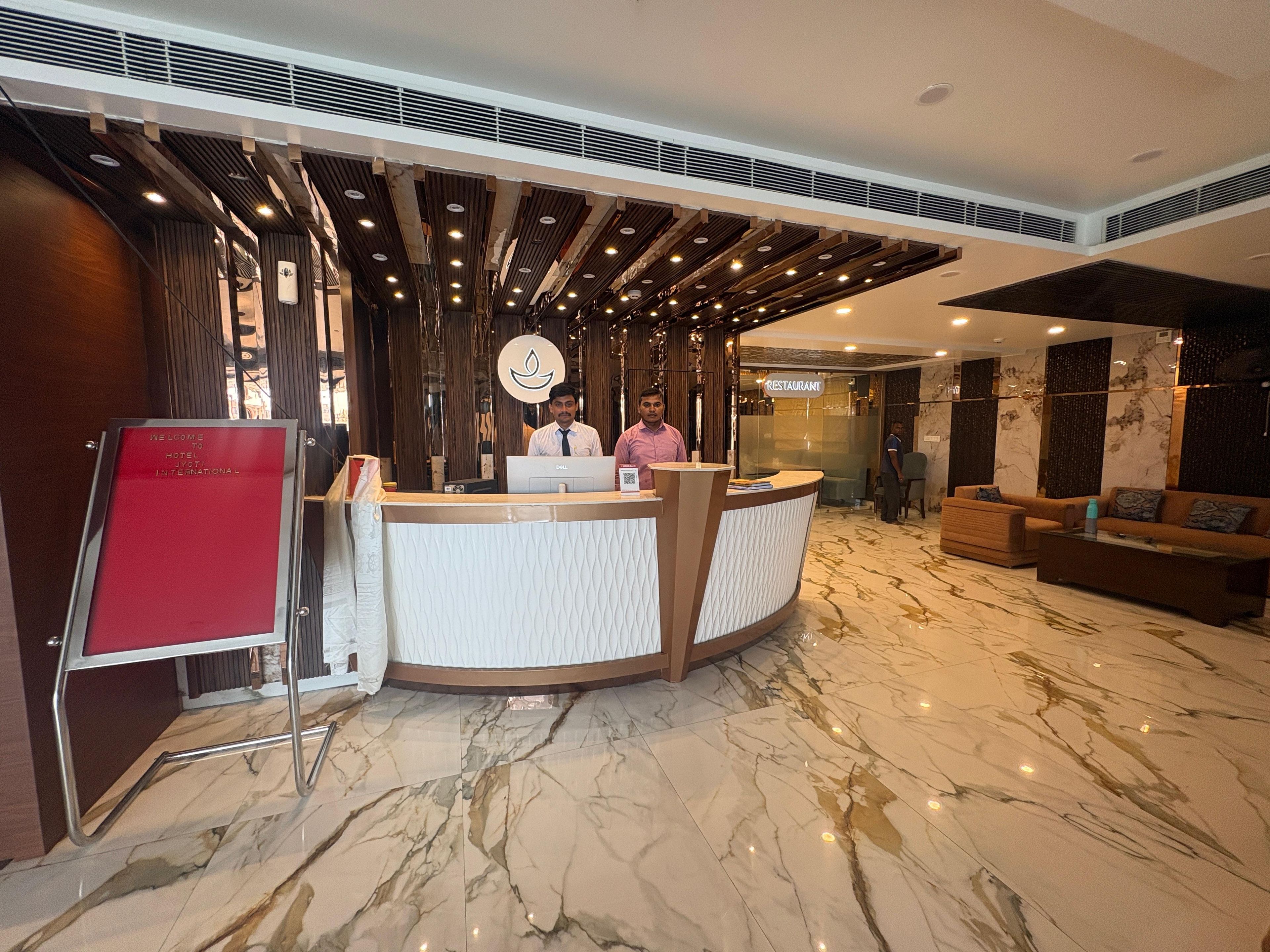 Zona de recepción de hotel con suelo de mármol, mostrador de recepción y un cartel que indica un restaurante.