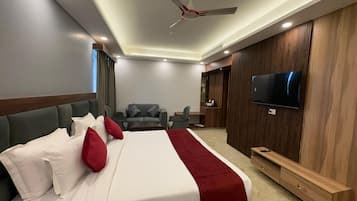 Una habitación de hotel moderna con una cama grande, un área de descanso con sofá y sillones, un televisor montado en la pared y un ventilador de techo.