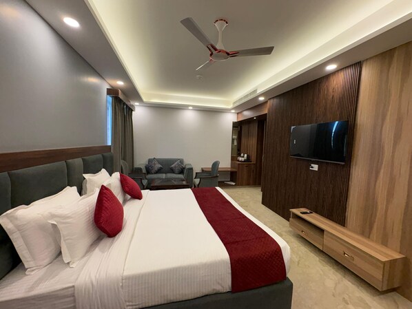 A modern hotel room with a large bed, a seating area with a sofa and armchairs, a television mounted on the wall, and a ceiling fan.