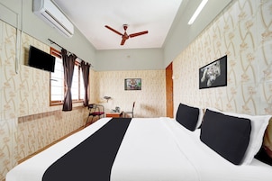A hotel room with a bed, a ceiling fan, a flat-screen TV, a desk with a chair, and a window with curtains.