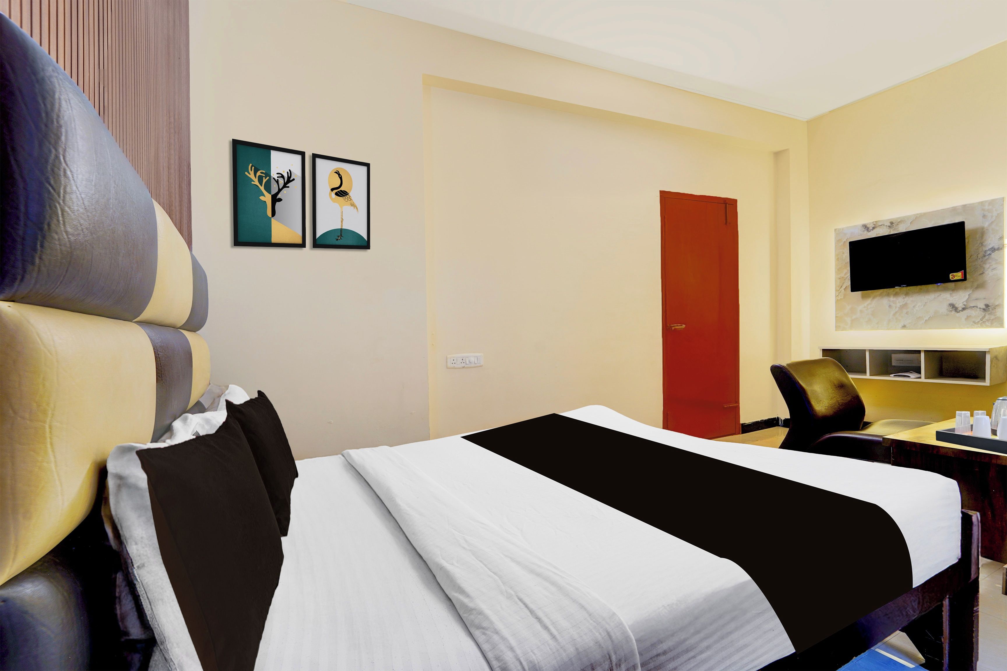 Quarto de hotel com cama, escrivaninha, cadeira, televisão e duas imagens emolduradas na parede.