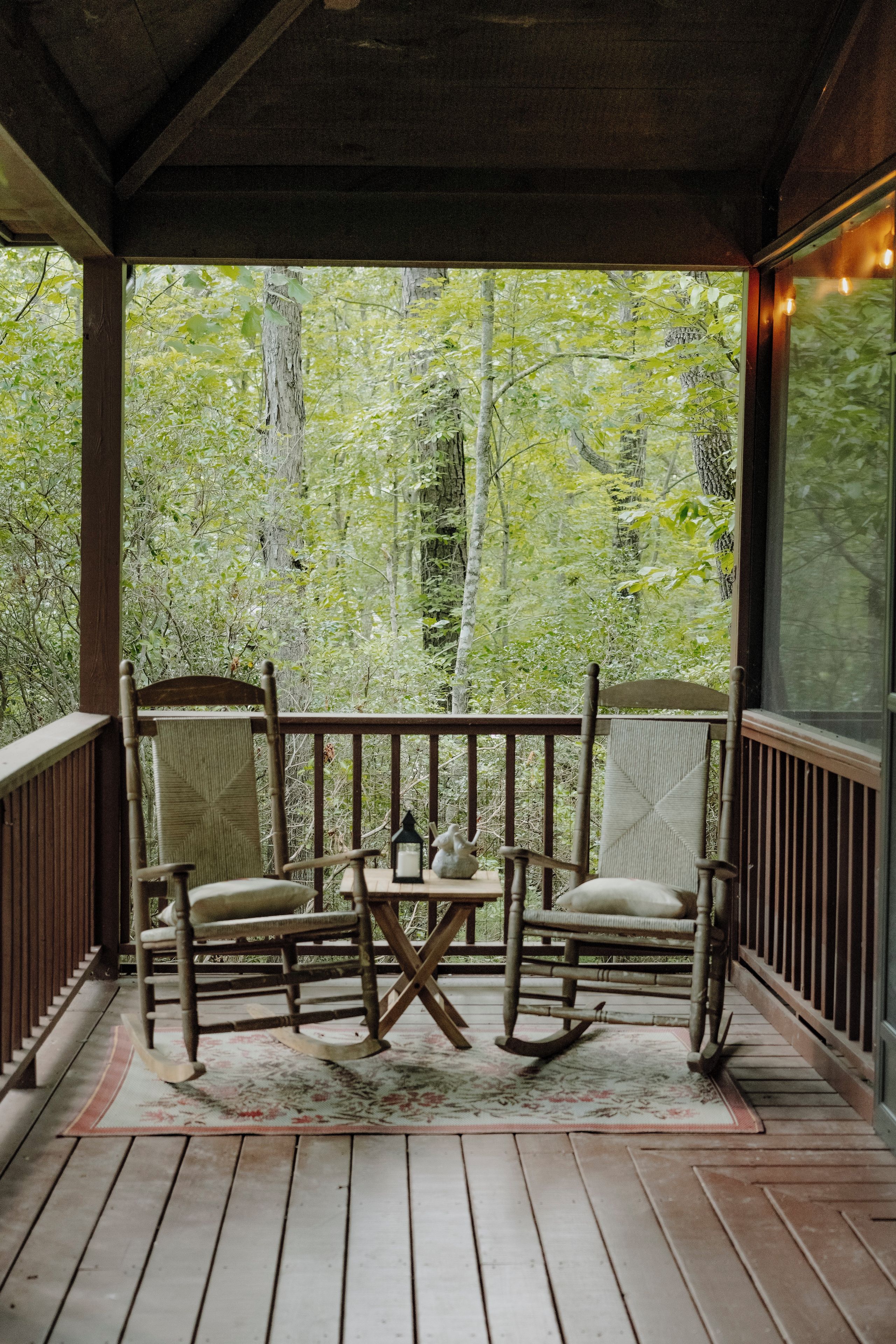 A wooden porch with two rocking chairs and a small table, overlooking a forested area.