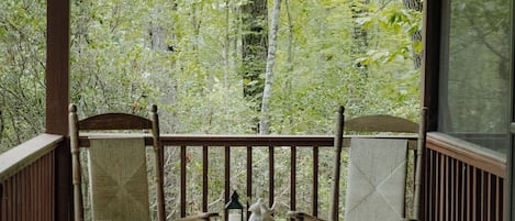 A wooden porch with two rocking chairs and a small table, overlooking a forested area.