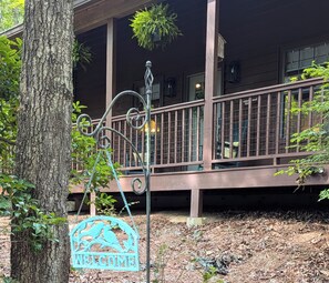 A wooden porch with a metal railing, a welcome sign, and hanging plants. - Rest & Unwind at Mountain Laurel Retreat 3-BR cabin in charming Sautee Nacoochee (Sautee Nacoochee)
