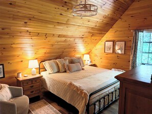 A cozy bedroom with a wooden ceiling, a bed with a quilt, bedside lamps, a nightstand, a chair, and a window with curtains. - Rest & Unwind at Mountain Laurel Retreat 3-BR cabin in charming Sautee Nacoochee (Sautee Nacoochee)