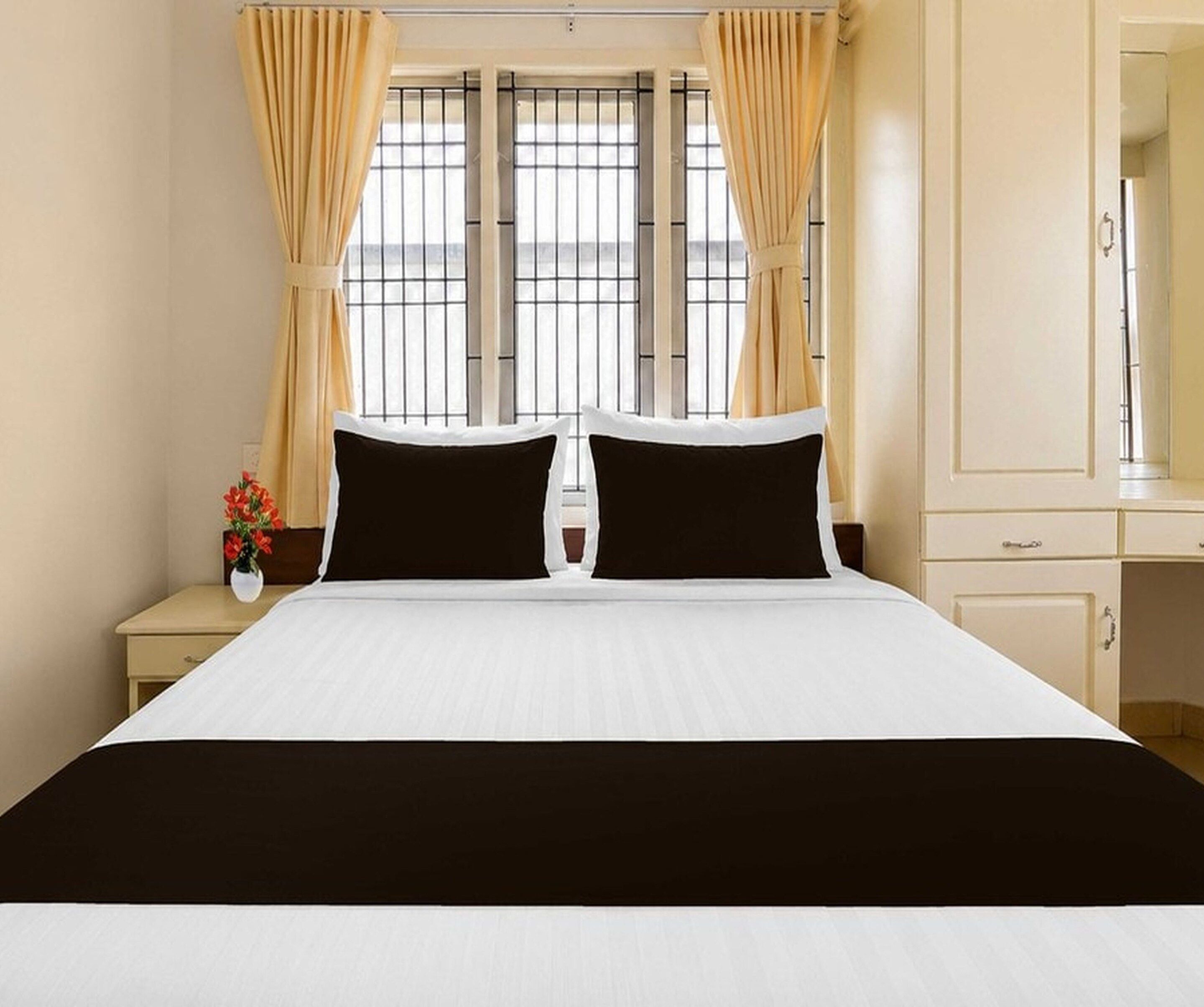 A hotel room with a large bed, two black pillows, a white bedspread, a nightstand with a vase of flowers, and a window with curtains.