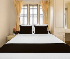 A hotel room with a large bed, two black pillows, a white bedspread, a nightstand with a vase of flowers, and a window with curtains. - Deluxe Room Sba's Park Inn (Coimbatore)
