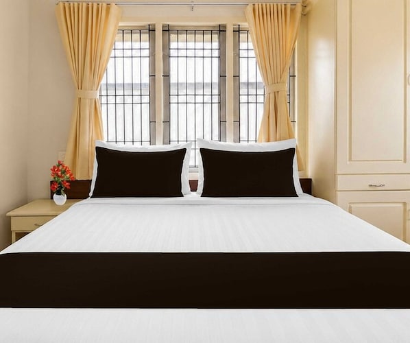 A hotel room with a large bed, two black pillows, a white bedspread, and beige curtains.