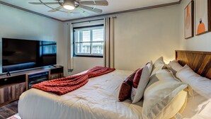 Quarto de hotel com uma cama, televisão, ventoinha no teto e janela com persianas.
