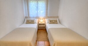 Una habitación pequeña y acogedora con dos camas individuales, una mesita de noche con lámpara y una ventana con cortinas traslúcidas.