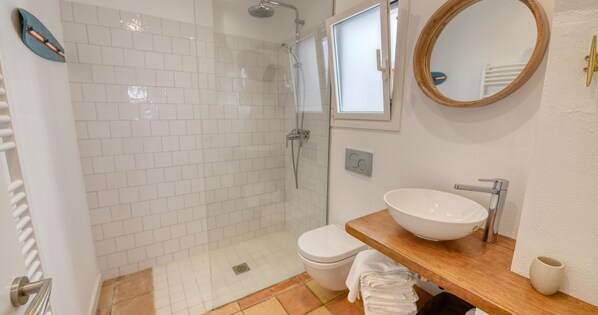A bathroom with a glass-enclosed shower, a white toilet, a wooden vanity with a white sink, and a round mirror. - Rustic-modern style apartment with sea views (Calella de Palafrugell)
