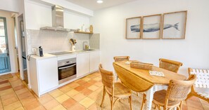 A modern kitchen with white cabinetry, a built-in oven, and a dining area with a wooden table and wicker chairs. - Rustic-modern style apartment with sea views (Calella de Palafrugell)
