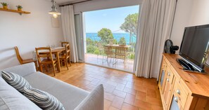 Een woonkamer met een bank, televisie, houten eettafel, stoelen en een balkon met uitzicht op zee.