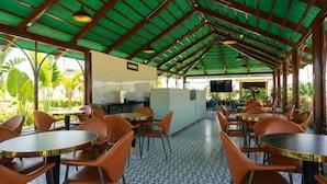 Um espaço de refeições interno com mesas redondas e cadeiras laranjas, piso ladrilhado e estrutura de teto verde.