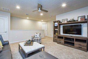 A living room with a sofa, ottoman, entertainment center, and ceiling fan. - 5BR Mario-themed home near Disney | Pool & Spa (Kissimmee)