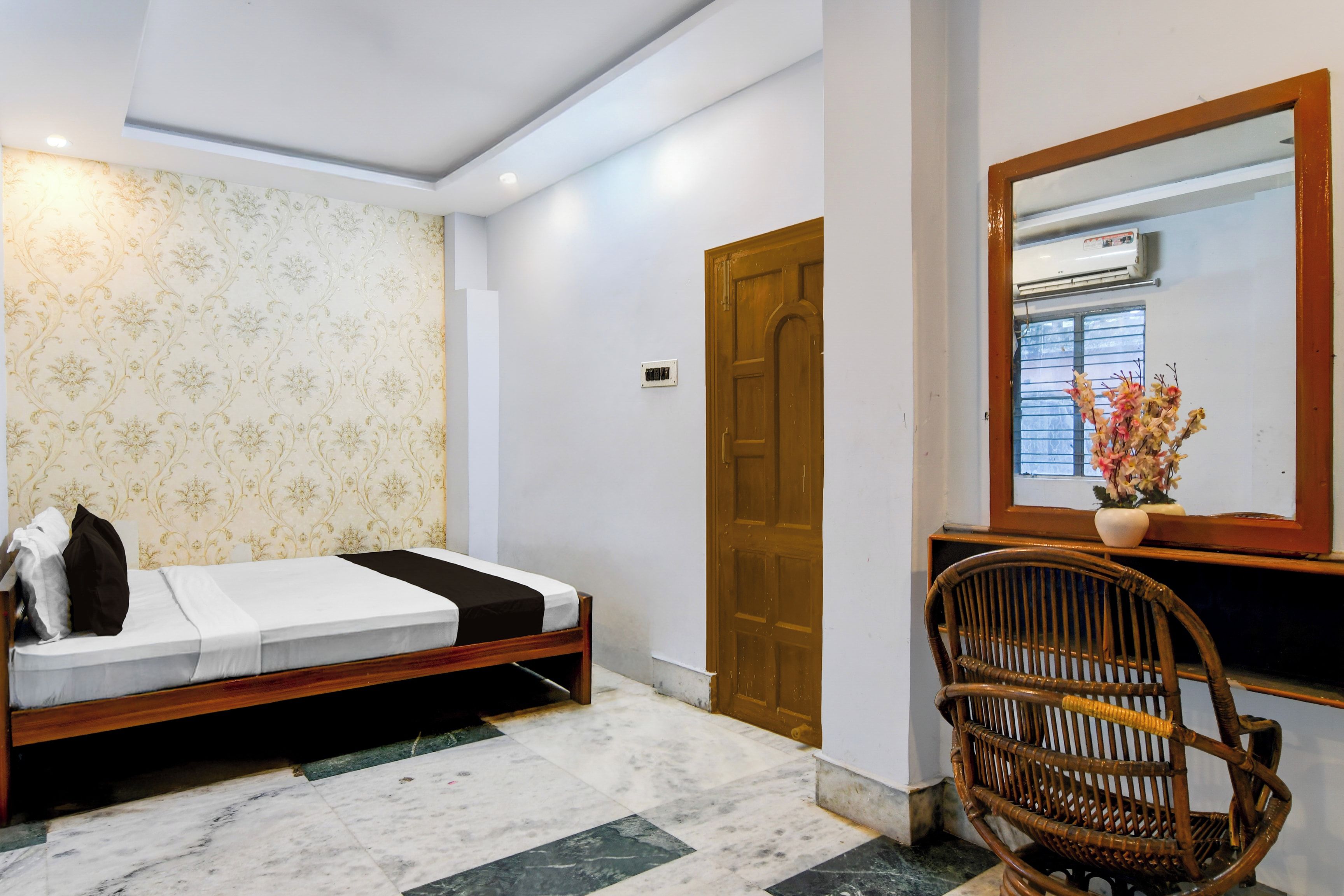 Une chambre d’hôtel avec un lit, une porte en bois, un miroir, une chaise en osier et un vase rempli de fleurs.