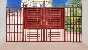 A red metal gate with vertical bars and a central latch, leading to a building with a white facade and blue windows.