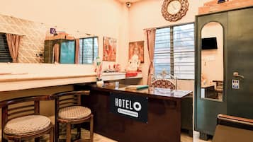 A hotel reception desk with a sign indicating it is company-serviced. There are two wooden chairs with patterned cushions in front of the desk.