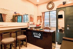 A hotel reception desk with a sign indicating it is company-serviced. There are two wooden chairs with patterned cushions in front of the desk.