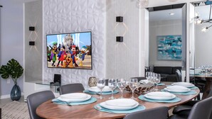 Ein moderner Essbereich mit einem großen Flachbildfernseher an der Wand, einem runden Holztisch, der für ein Essen gedeckt ist, und einer großen Topfpflanze.