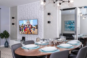 Un área de comedor moderna con un televisor de pantalla plana grande montado en la pared, una mesa redonda de madera preparada para comer y una planta grande en maceta.