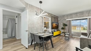 A modern living room with a dining area, a yellow accent wall, a large window with a view, and a grey sofa. - 1BR ADA apartment | 5 min to Epic Universe (Orlando)