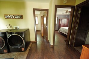 A laundry area with a washing machine and dryer, a wooden door with a 'welcome' sign, a hallway leading to a bedroom with a bed and a wardrobe. - Riverfront Property Near Downtown Pella with Game Room, Fire Pit, and Yard Games (Pella)