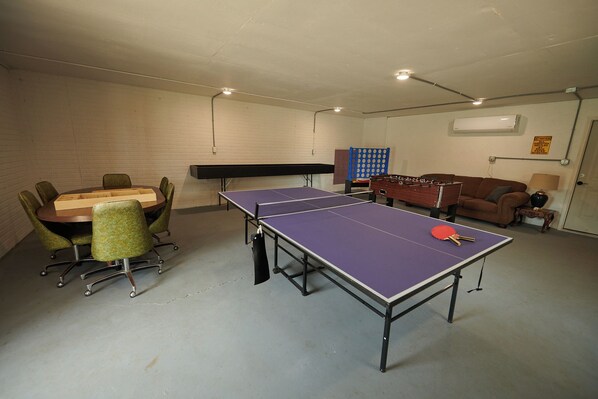 A room with a ping pong table, a foosball table, a table tennis table, and a sofa.