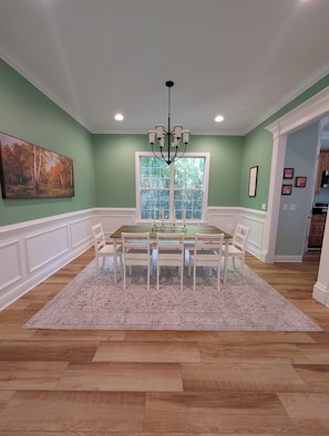 A dining room with a wooden table, white chairs, a chandelier, and a large window with a view of trees.