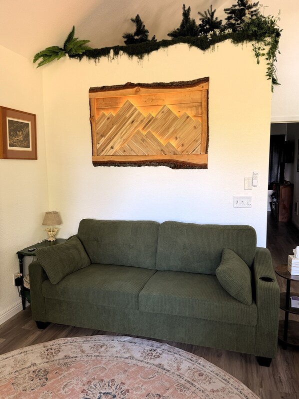 A green sofa in a room with a wooden wall decoration, a framed picture, and a lamp on a side table.