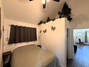 Un dormitorio con cama, mesita de noche, lámpara de pared, ventana con cortinas y puerta que da a otra habitación.
