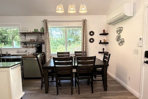 A dining area with a table and chairs, a kitchen with shelves, and a window with curtains.