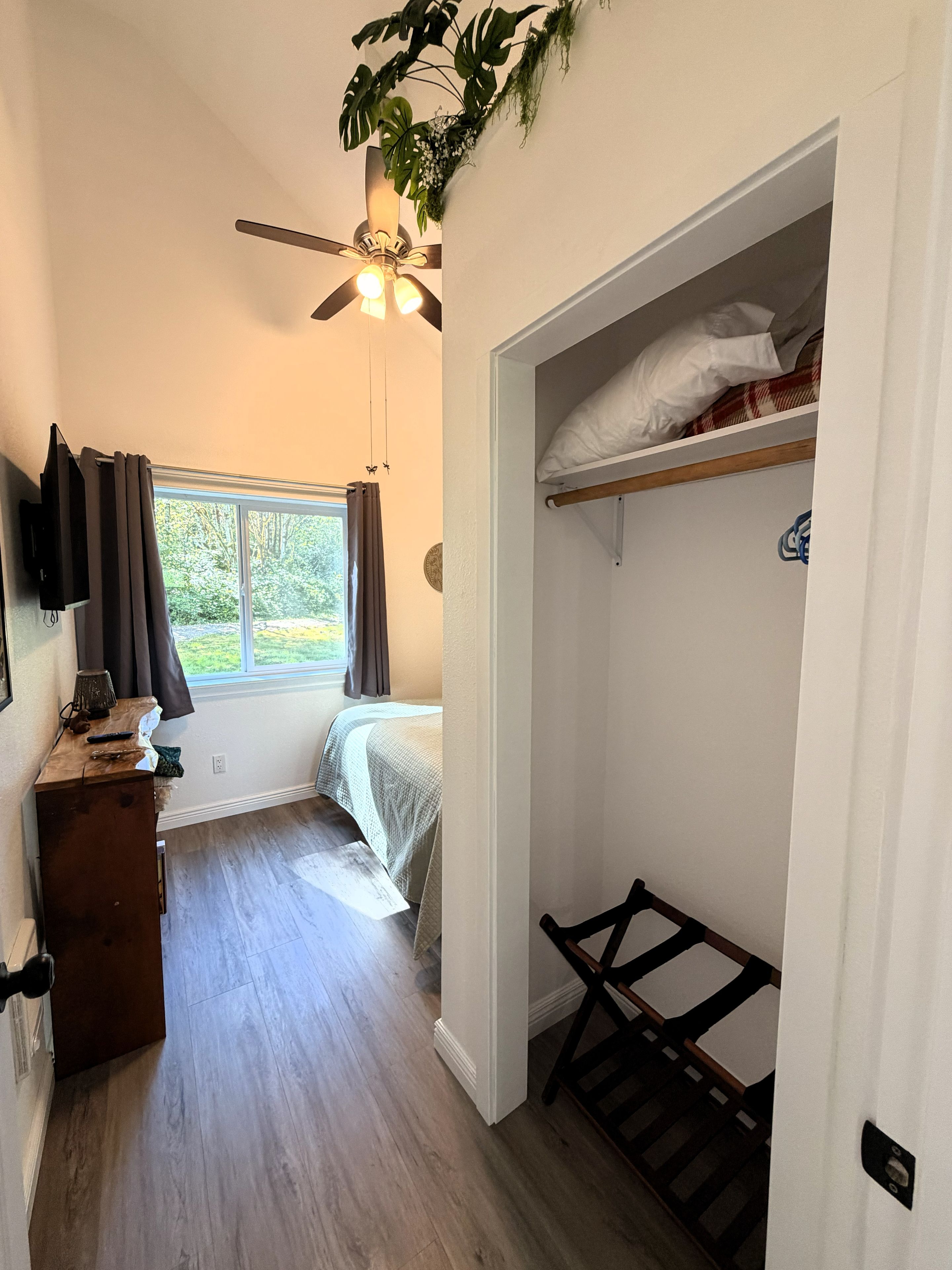 A bedroom with a bed, a ceiling fan, a window with curtains, a wooden dresser, and a closet with a shelf.