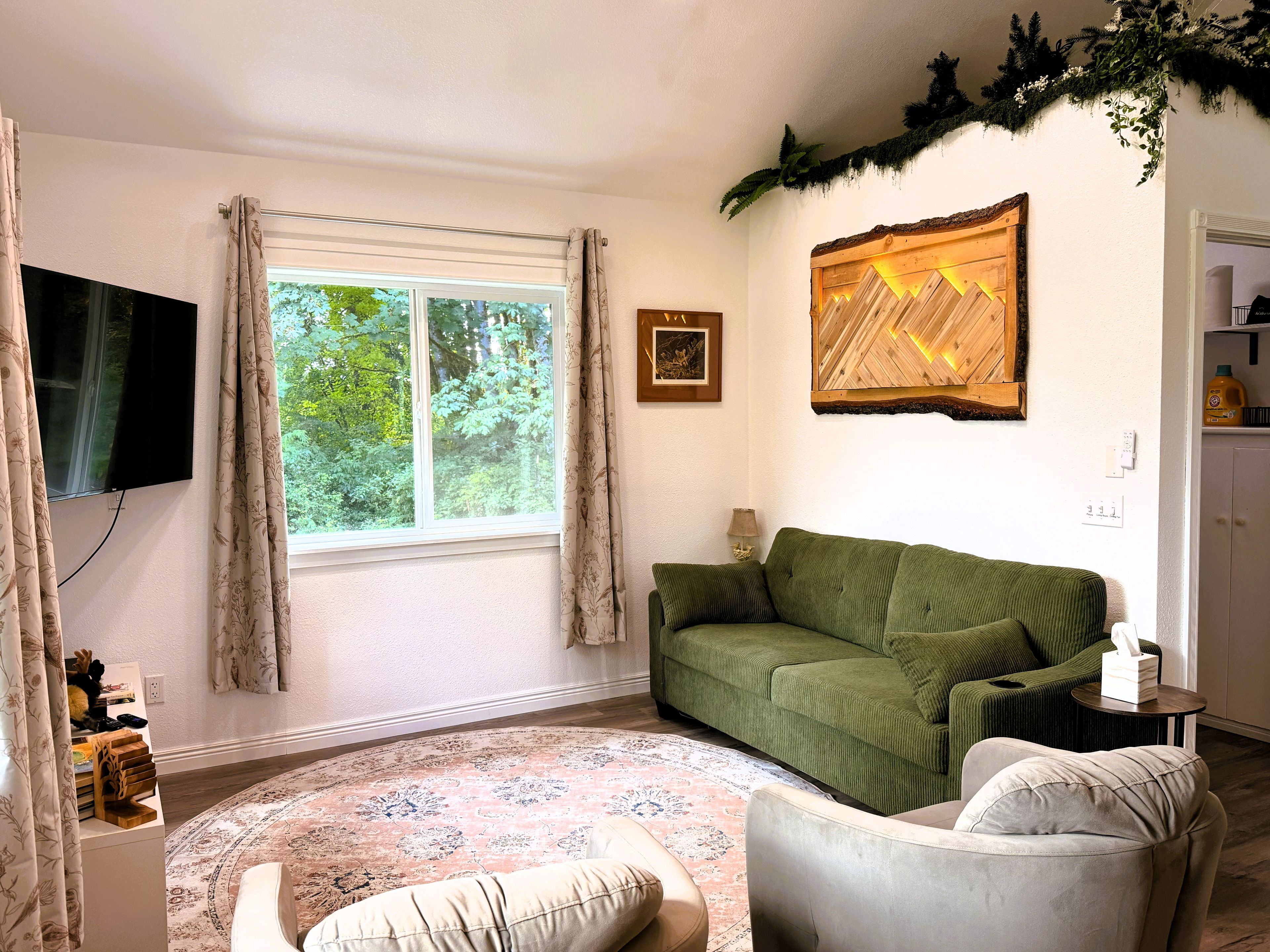 A living room with a green sofa, a grey armchair, a patterned rug, a flat-screen TV, and a window with curtains.